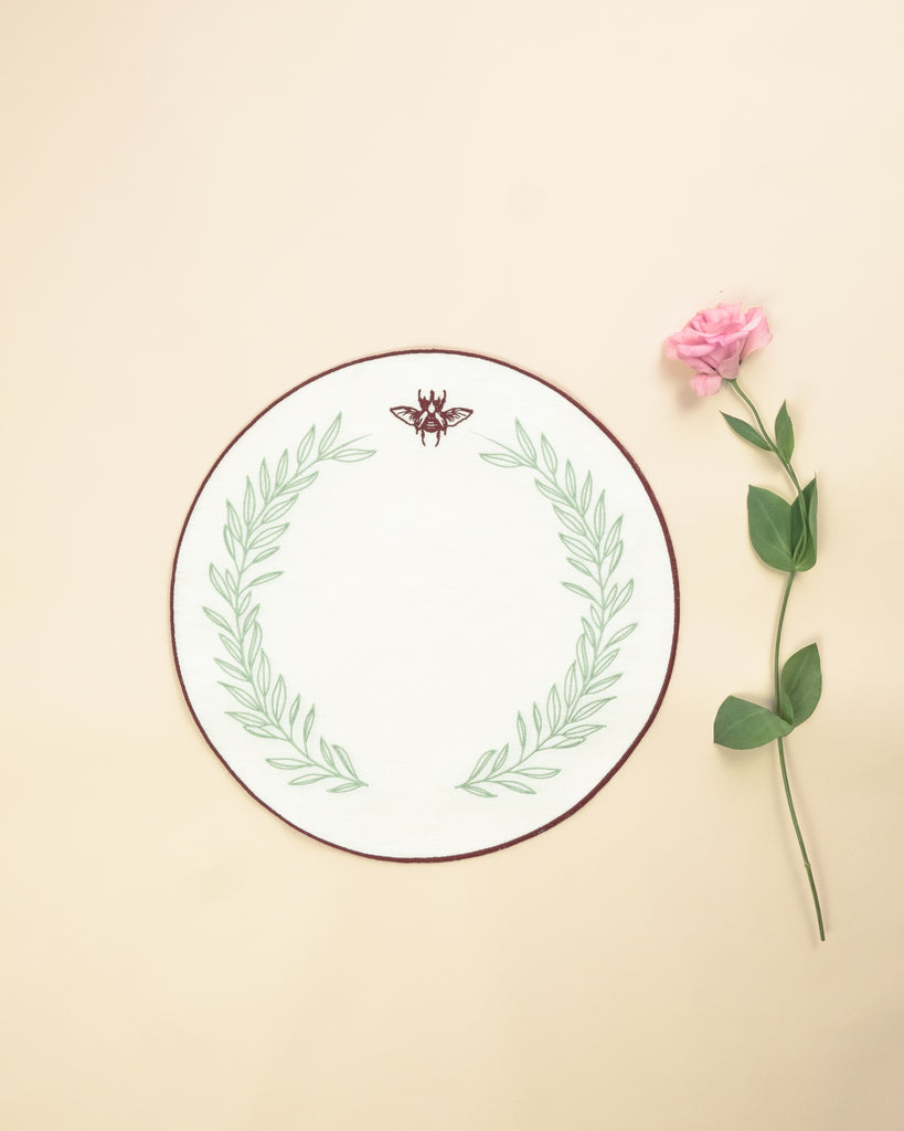 Round placemat in white with embroidered bordeaux border and a bordeaux scarab and green foliage embroidered