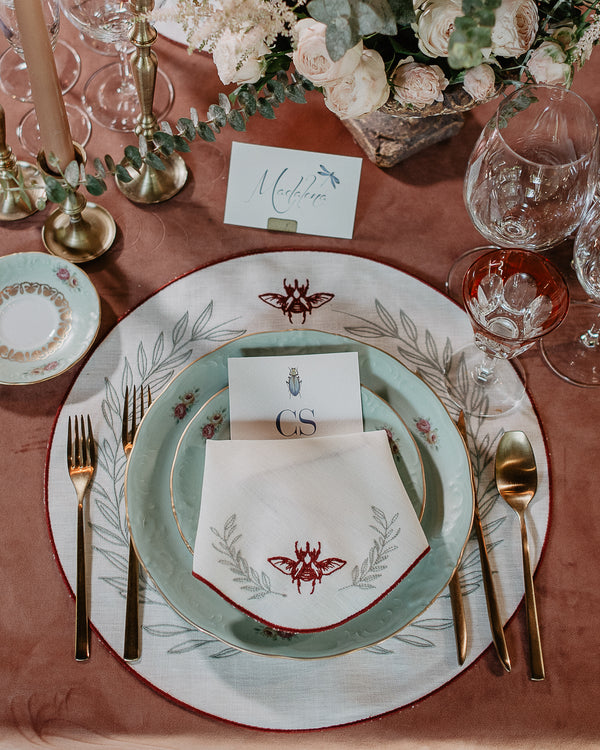 Festive table setting with matching napkin and placemat, both with bordeaux embroidered borders, a bordeaux embroidered scarab and embroidered green foliage