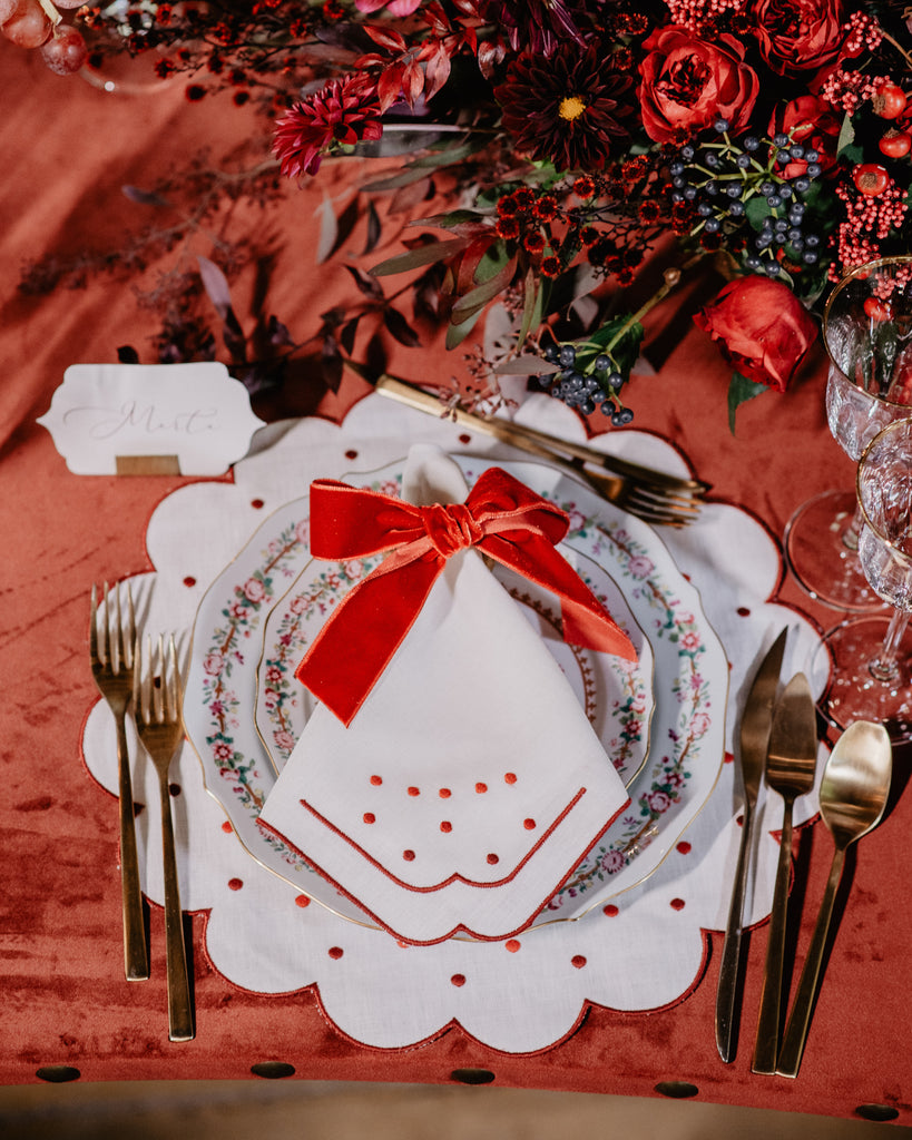 White napkin with bordeaux embroidered borders and details, envolved in a red ribbon, stands on top of a flower-themed ceramic plate placed on top of a flower-shaped placemat matching the napkin embroideries